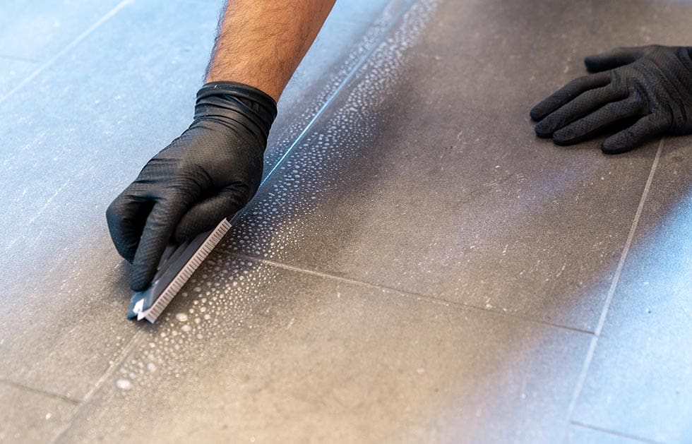 close up of professional cleaner cleaning grout with a brush blade and foamy soap on a gray tiled bathroom floor